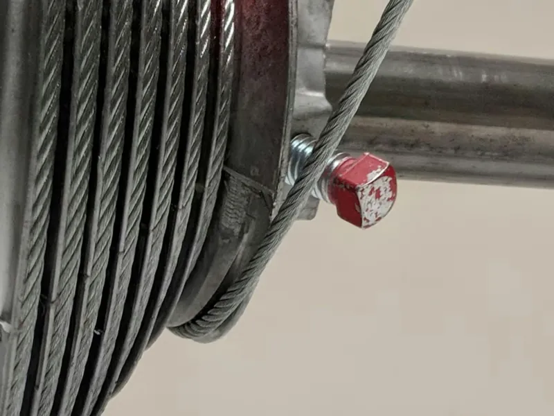 Close view of a steel lift cable wound on a cable drum at the end of a torsion shaft, above the top corner of a white sectional garage door.