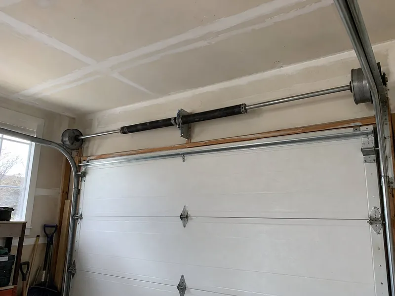 Inside a garage, a black torsion spring is wound on a steel shaft above a closed white sectional door, with cable drums at both ends and the steel tracks curving overhead.